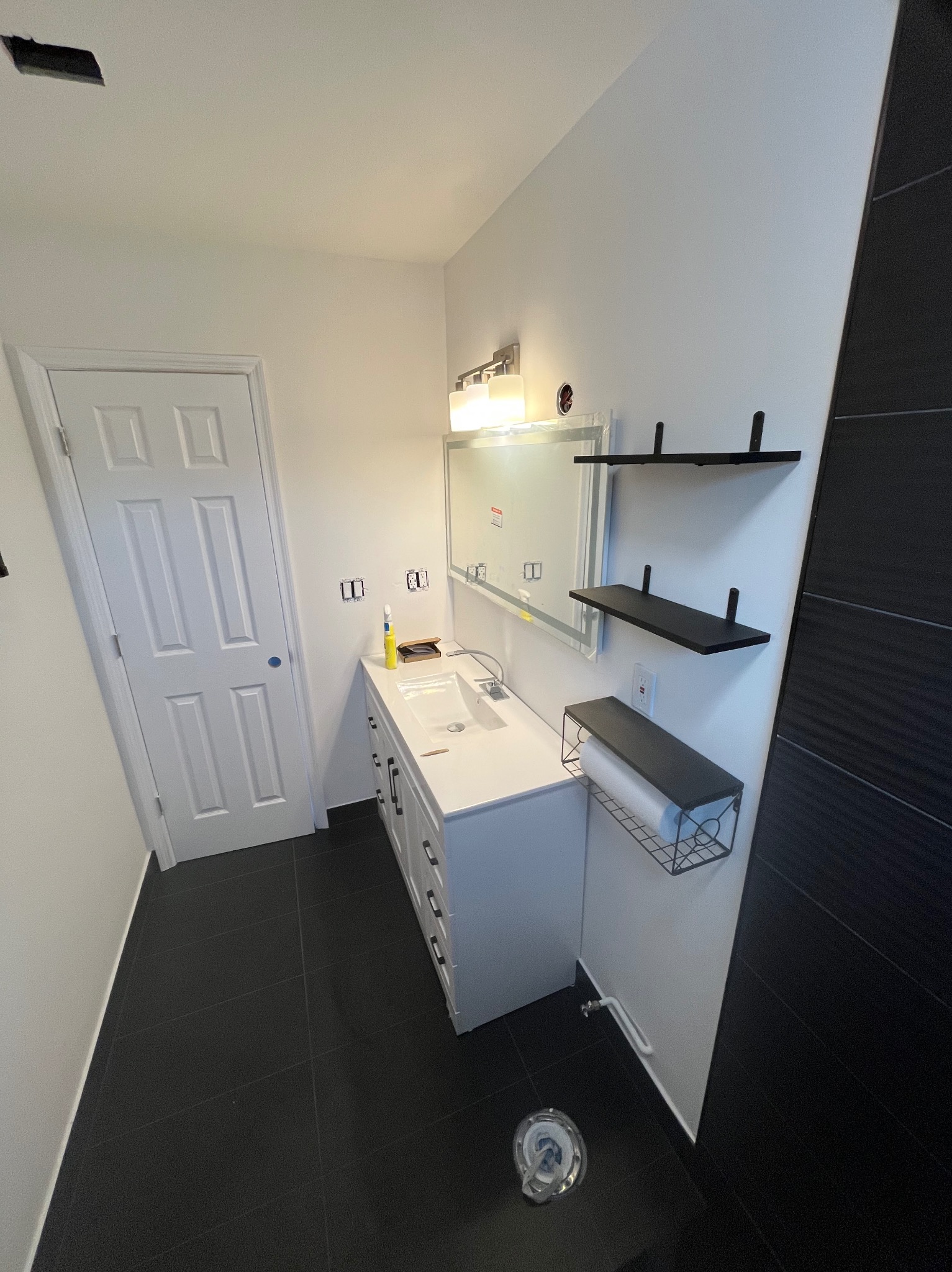 Nowoczesna łazienka po remoncie: biała szafka z umywalką, podświetlane lustro, czarne półki i ciemna podłoga. Widoczne detale wykończenia i instalacje.