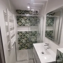 Nowoczesna łazienka z kabiną prysznicową, białą umywalką i grzejnikiem drabinkowym. Uwagę przyciągają płytki z motywem tropikalnych liści.