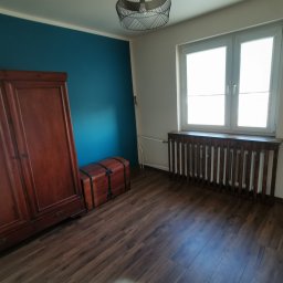 Paweł Chmiel FUTURE - Pokój z drewnianą podłogą, szafą i skrzynią. Ściana pomalowana na niebiesko, okno z drewnianą obudową grzejnika. Widok na wnętrze po wykończeniu.
