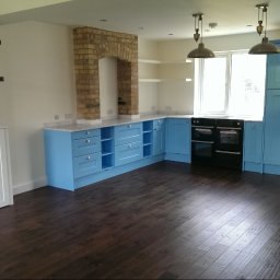 Paweł Chmiel FUTURE - Jasna kuchnia z błękitnymi szafkami i ciemną podłogą. Ceglany komin nad blatem. Dwa wiszące światła. Biała szafka w zabudowie. Nowoczesny design.