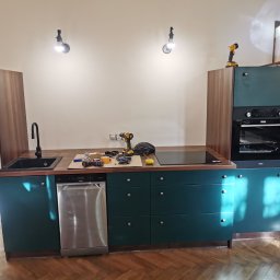 Paweł Chmiel FUTURE - Montaż zielonej kuchni z drewnianym blatem i sprzętem AGD. Widoczne narzędzia pracy na blacie. Parkiet na podłodze. Dwie lampy oświetlają blat.