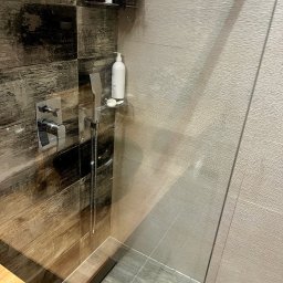 Czyste Wnętrze Magdalena Dykalska - Nowoczesna kabina prysznicowa z chromowaną armaturą, półką na kosmetyki i szklaną ścianą. Widoczne płytki imitujące drewno i odpływ liniowy.