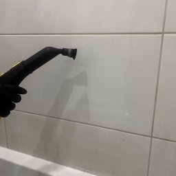 Czyste Wnętrze Magdalena Dykalska - Czyszczenie parowe płytek w łazience przy użyciu parownicy Kärcher, widoczna ręka w czarnej rękawicy, białe płytki i fragment wanny.