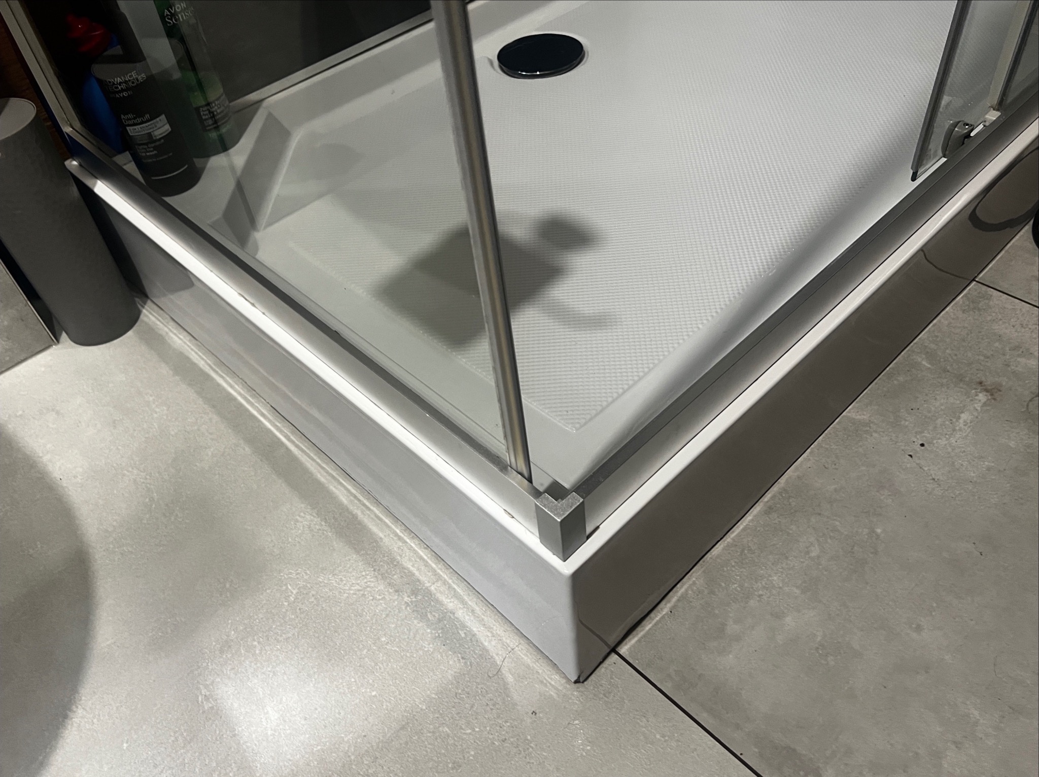 Biały brodzik kabiny prysznicowej z odpływem, widoczny fragment szklanej ścianki i kosmetyki na półce w tle, na szarych płytkach podłogowych.