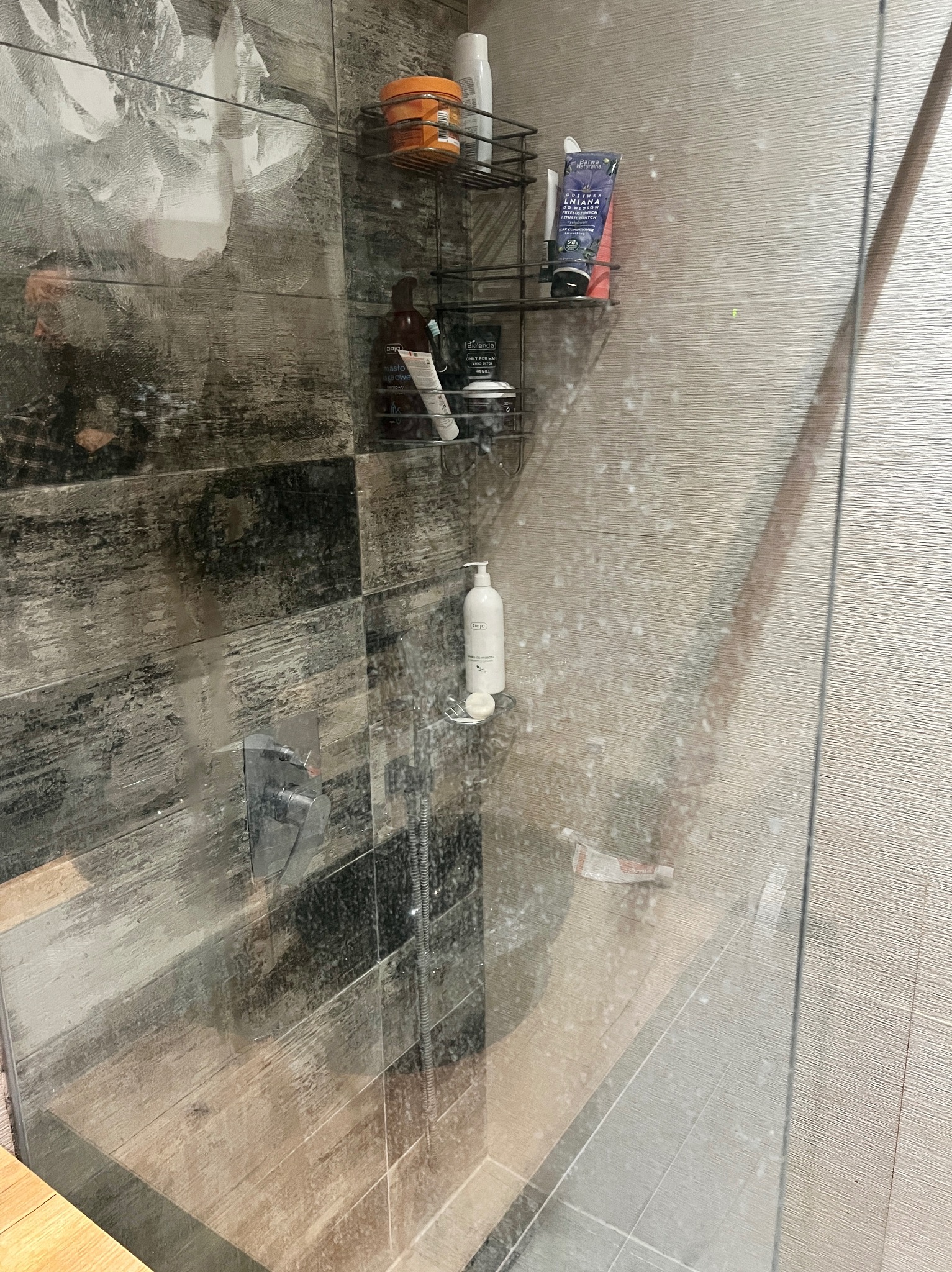 Zakamieniona kabina prysznicowa z półkami pełnymi kosmetyków. Widoczne ślady po wodzie na szklanej powierzchni i płytkach imitujących drewno.