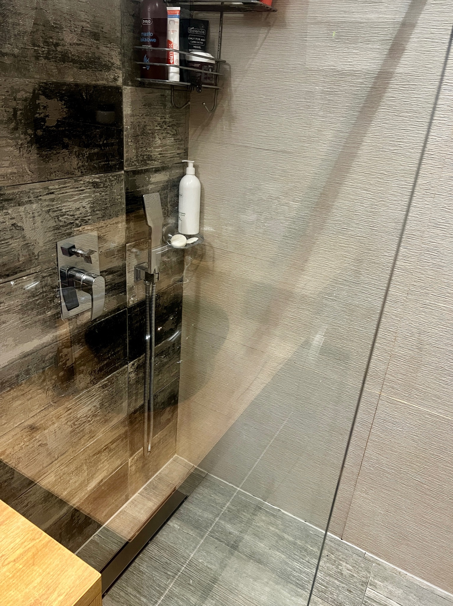 Nowoczesna kabina prysznicowa z chromowaną armaturą, półką na kosmetyki i szklaną ścianą. Widoczne płytki imitujące drewno i odpływ liniowy.