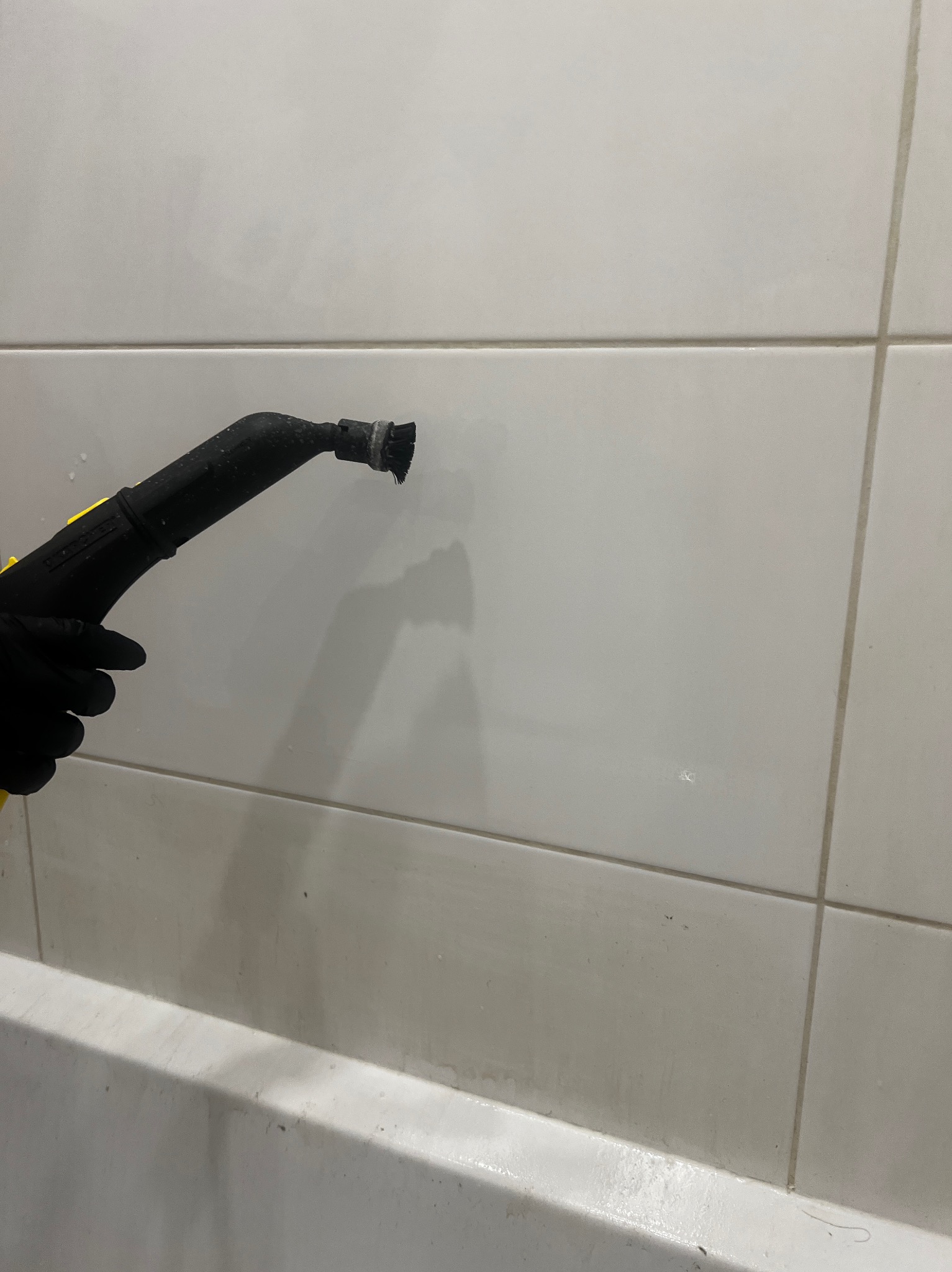 Czyszczenie parowe płytek w łazience przy użyciu parownicy Kärcher, widoczna ręka w czarnej rękawicy, białe płytki i fragment wanny.