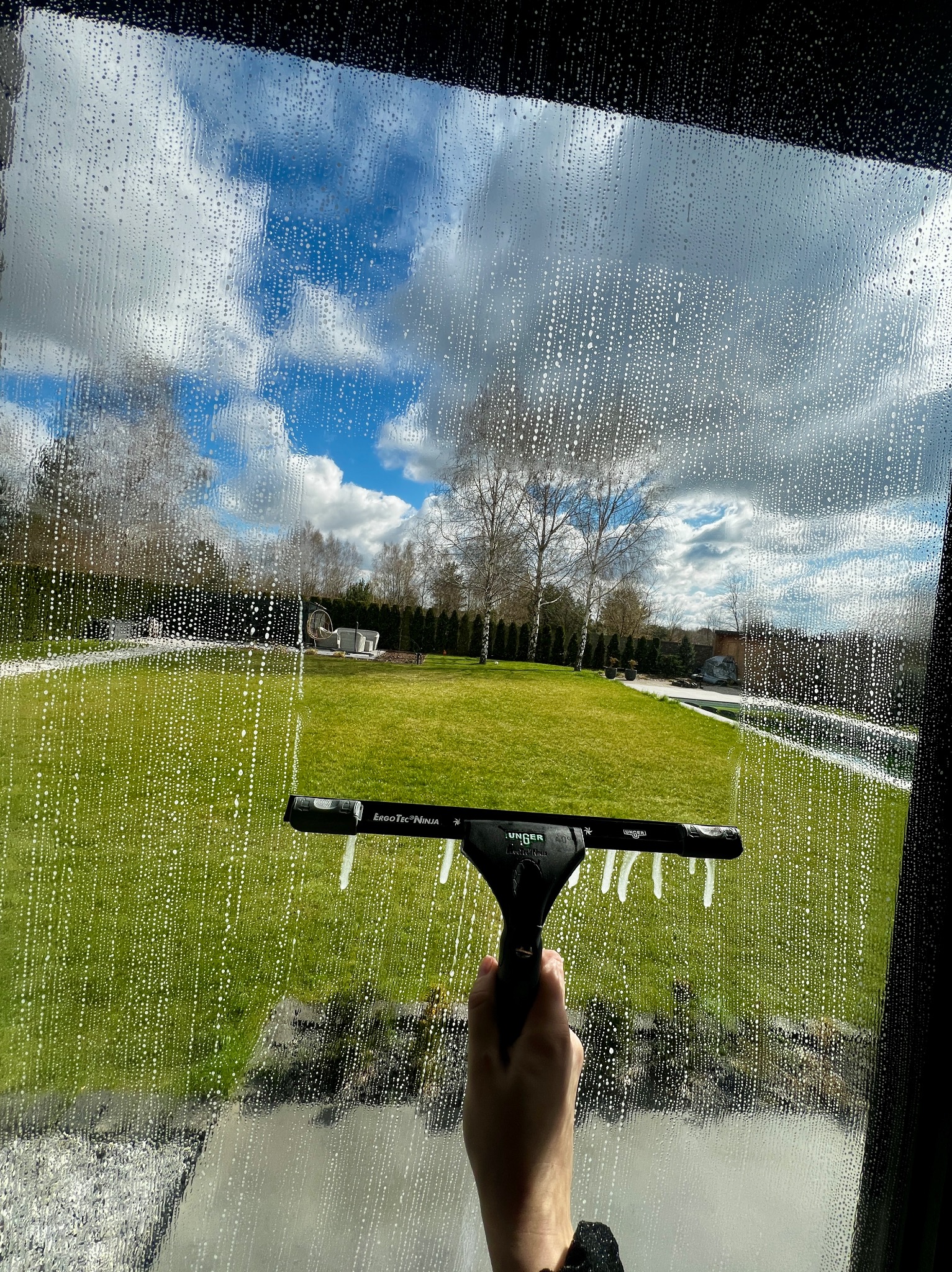 Mycie okna z widokiem na ogród. Na szybie piana i ślady po ściągaczce Unger ErgoTec Ninja, w tle trawnik, drzewa i meble ogrodowe.