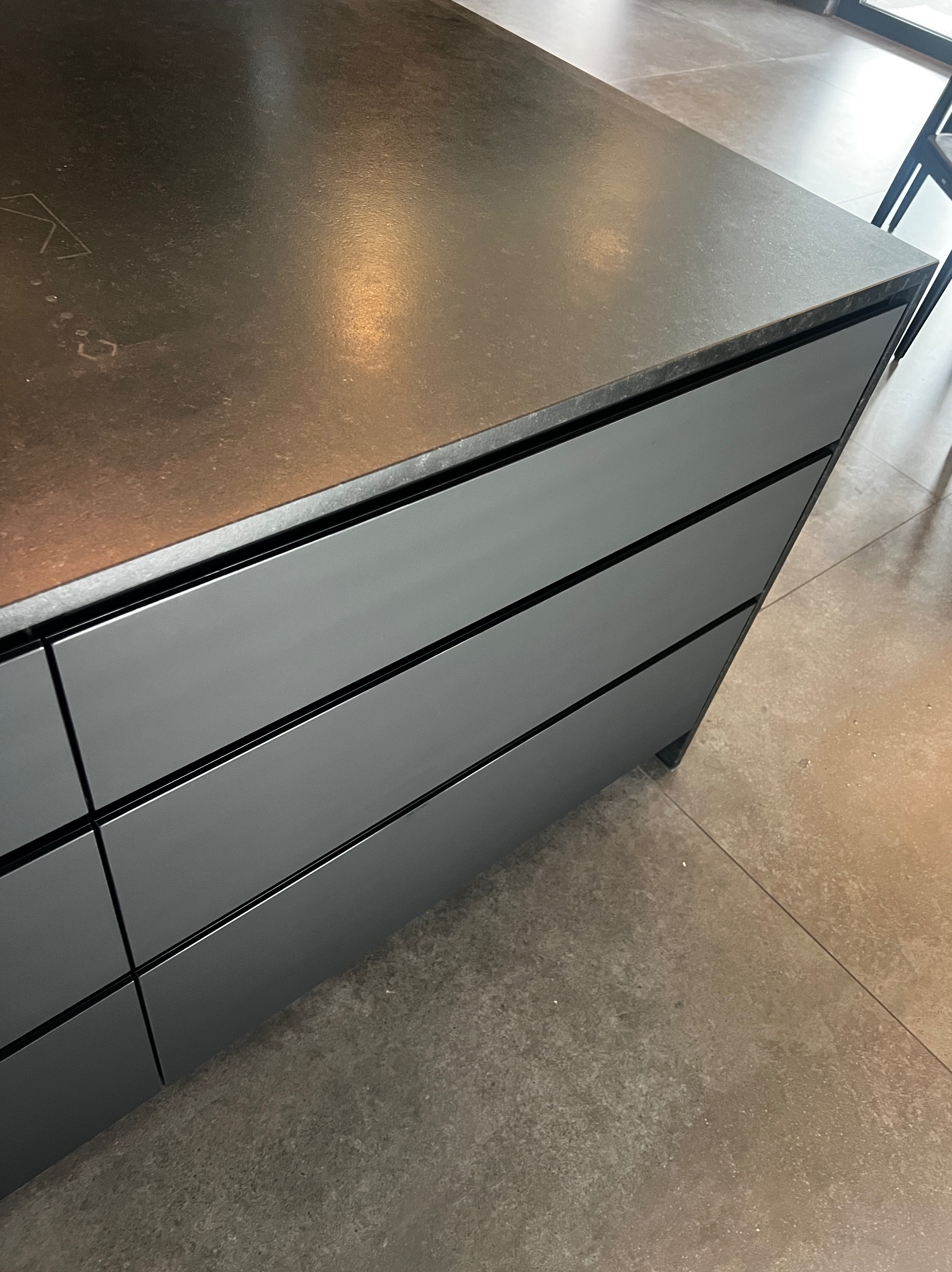 Nowoczesna wyspa kuchenna z szarymi frontami i blatem o teksturze kamienia, widoczna z bliska, z fragmentem podłogi z płytek w tle.