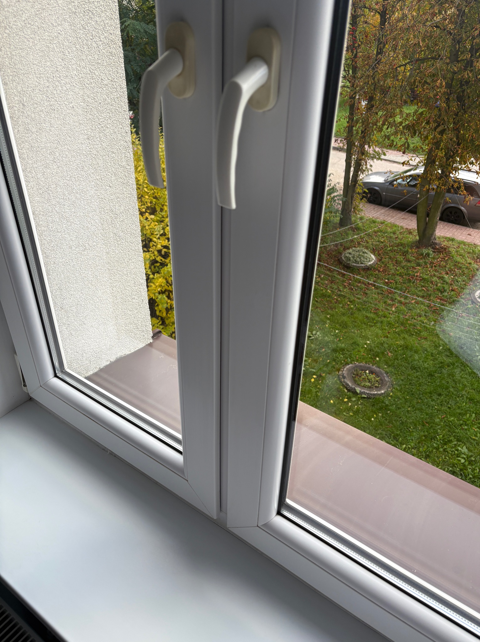 Białe, plastikowe okno z widokiem na zewnątrz. Widoczny fragment ściany budynku i trawnik z drzewami. Okno zamknięte, z widocznymi białymi klamkami.