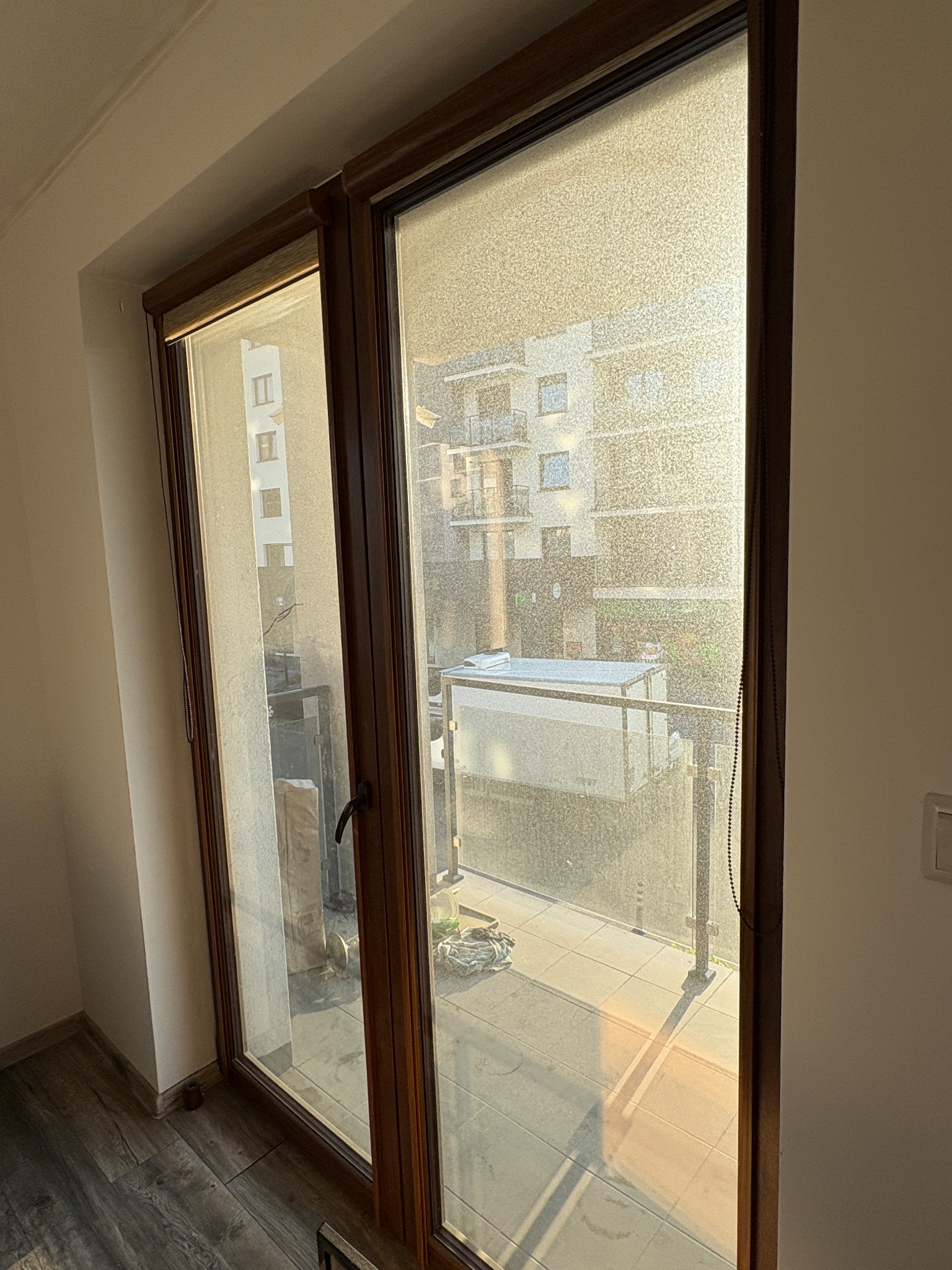 Bardzo brudne okno balkonowe z drewnianą ramą, widok na balkon i budynek mieszkalny. Silne zabrudzenia na szybie utrudniają widoczność.