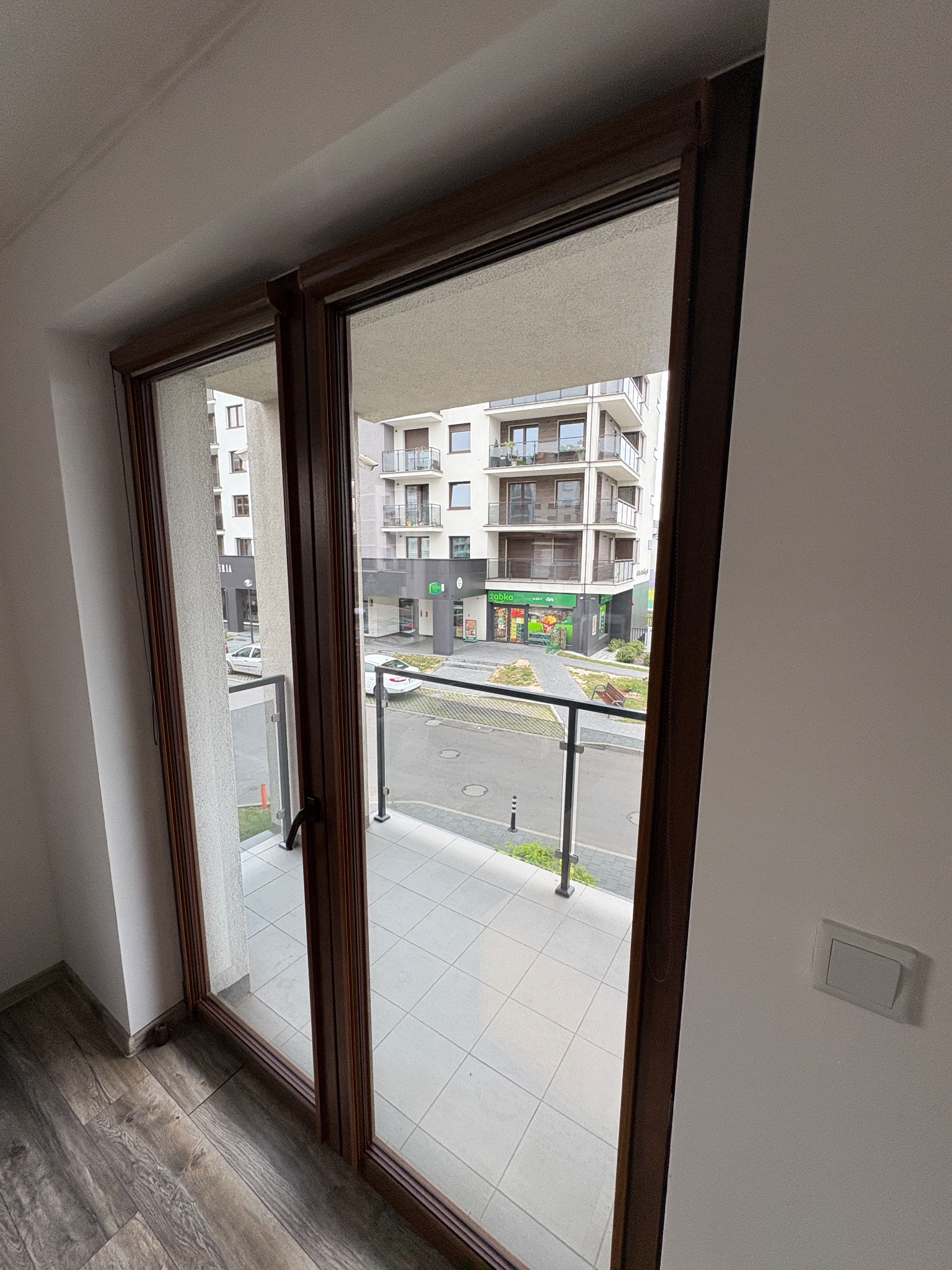 Widok z balkonu przez otwarte, dwuskrzydłowe drzwi balkonowe z brązową ramą. Na zewnątrz widoczny budynek mieszkalny i ulica.