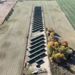 Widok z lotu ptaka na długi rząd paneli słonecznych umieszczonych na polu, otoczonych drzewami i polami uprawnymi w jesiennej scenerii.