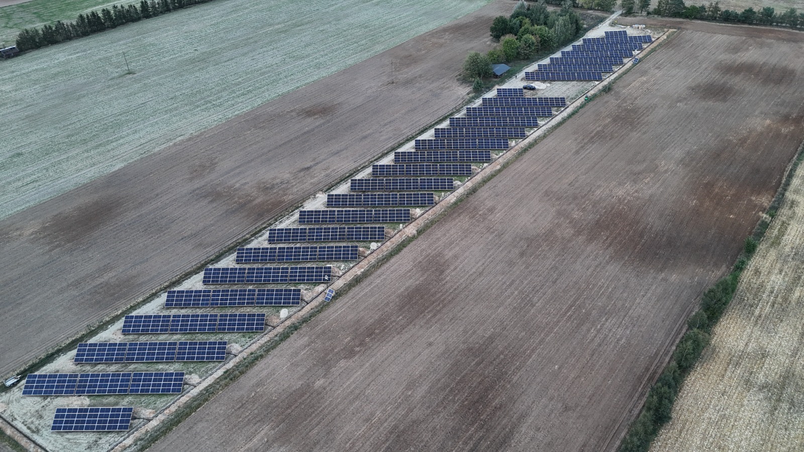 Widok z lotu ptaka na rozległą farmę paneli słonecznych, otoczoną polami w jesiennej kolorystyce, z widocznymi śladami po orce i niewielkim budynkiem gospodarczym w tle.
