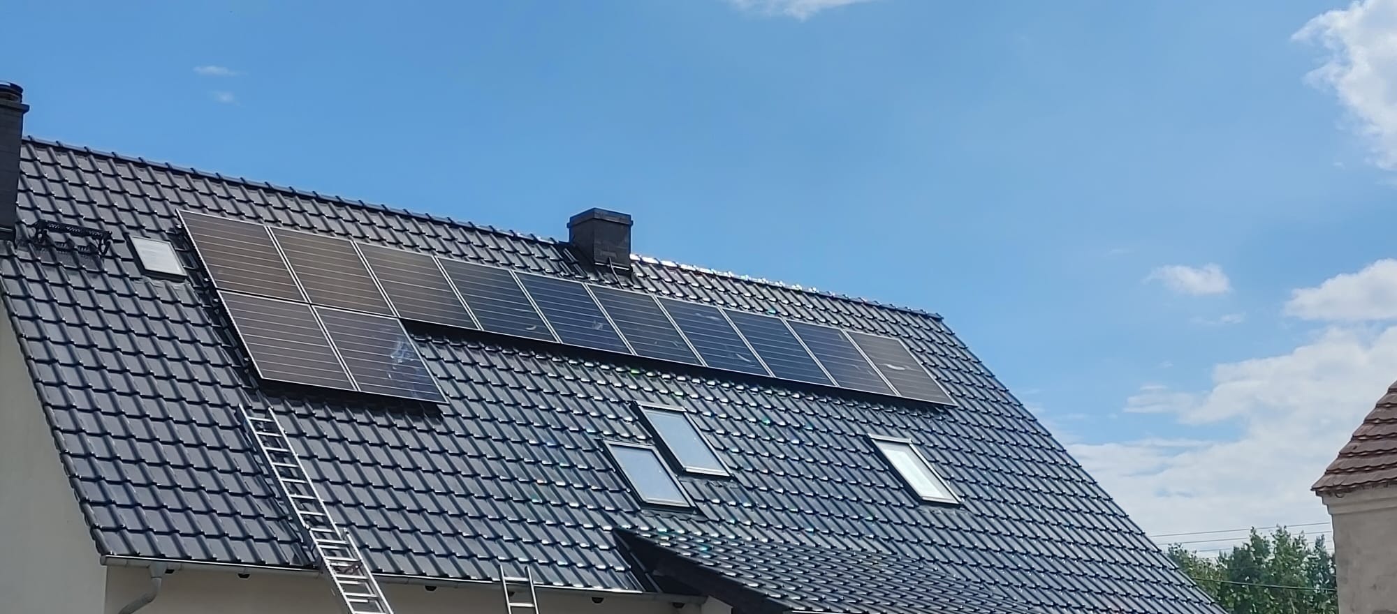 Ciemne panele słoneczne zamontowane na spadzistym dachu pokrytym ciemną dachówką, z widoczną drabiną opartą o ścianę budynku i oknami dachowymi, na tle błękitnego nieba z pojedynczymi chmurami.