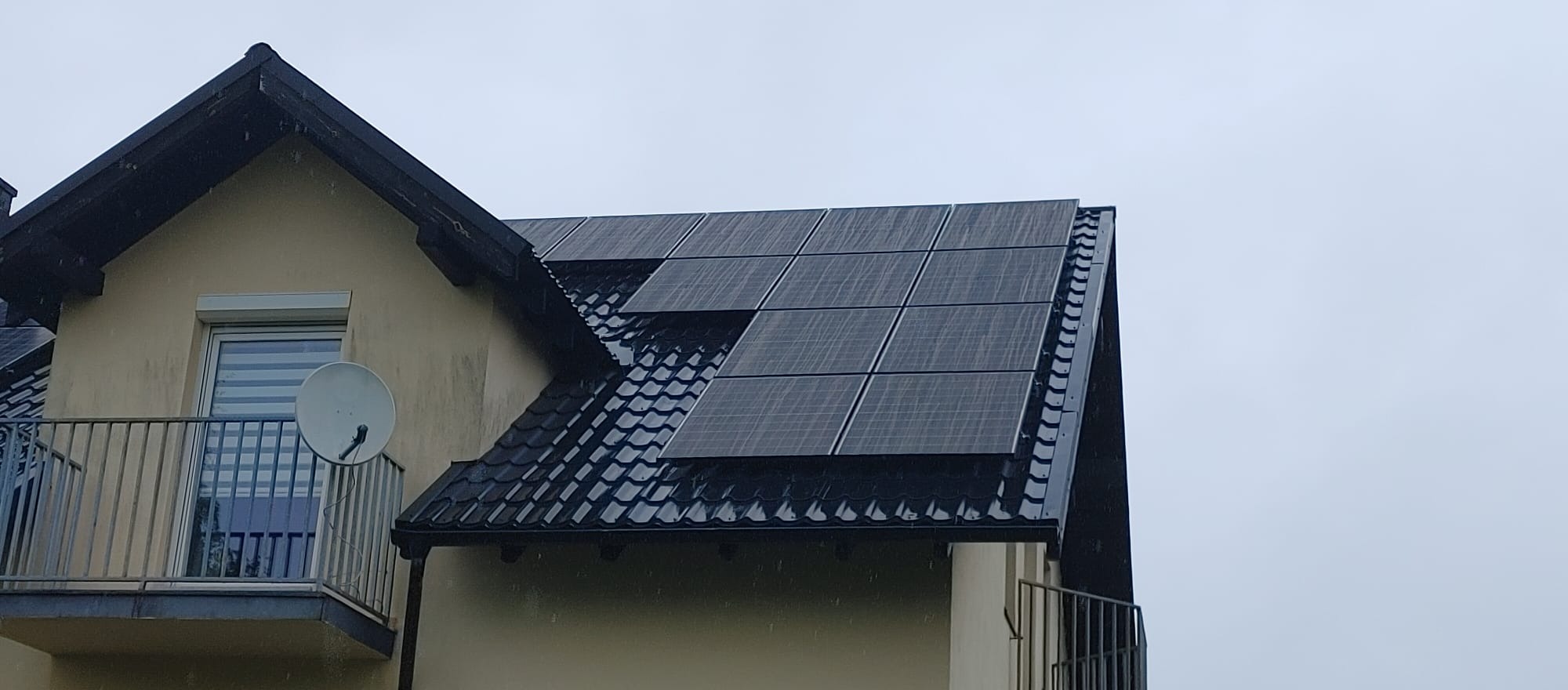 Czarny dach z panelami słonecznymi w deszczowy dzień, widoczny balkon z szarymi barierkami i satelitą.