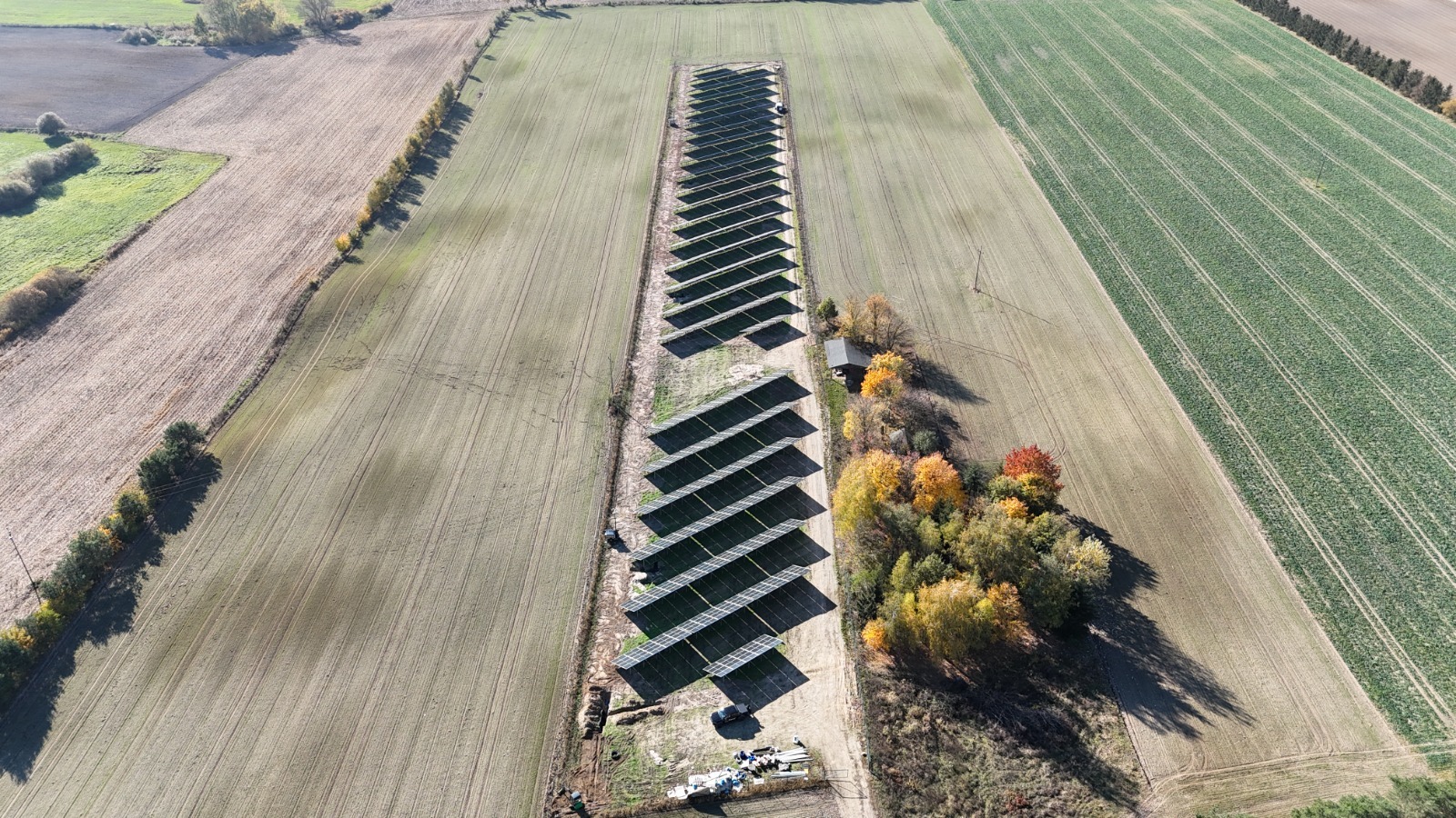 Widok z lotu ptaka na długi rząd paneli słonecznych umieszczonych na polu, otoczonych drzewami i polami uprawnymi w jesiennej scenerii.