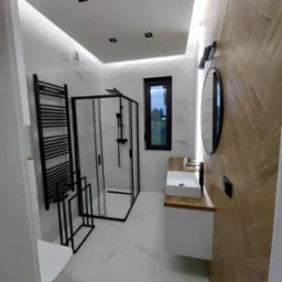 Nowoczesna łazienka z kabiną prysznicową typu walk-in, grzejnikiem drabinkowym i drewnopodobną ścianą ułożoną w jodełkę. Białe marmurowe płytki i podświetlany sufit.