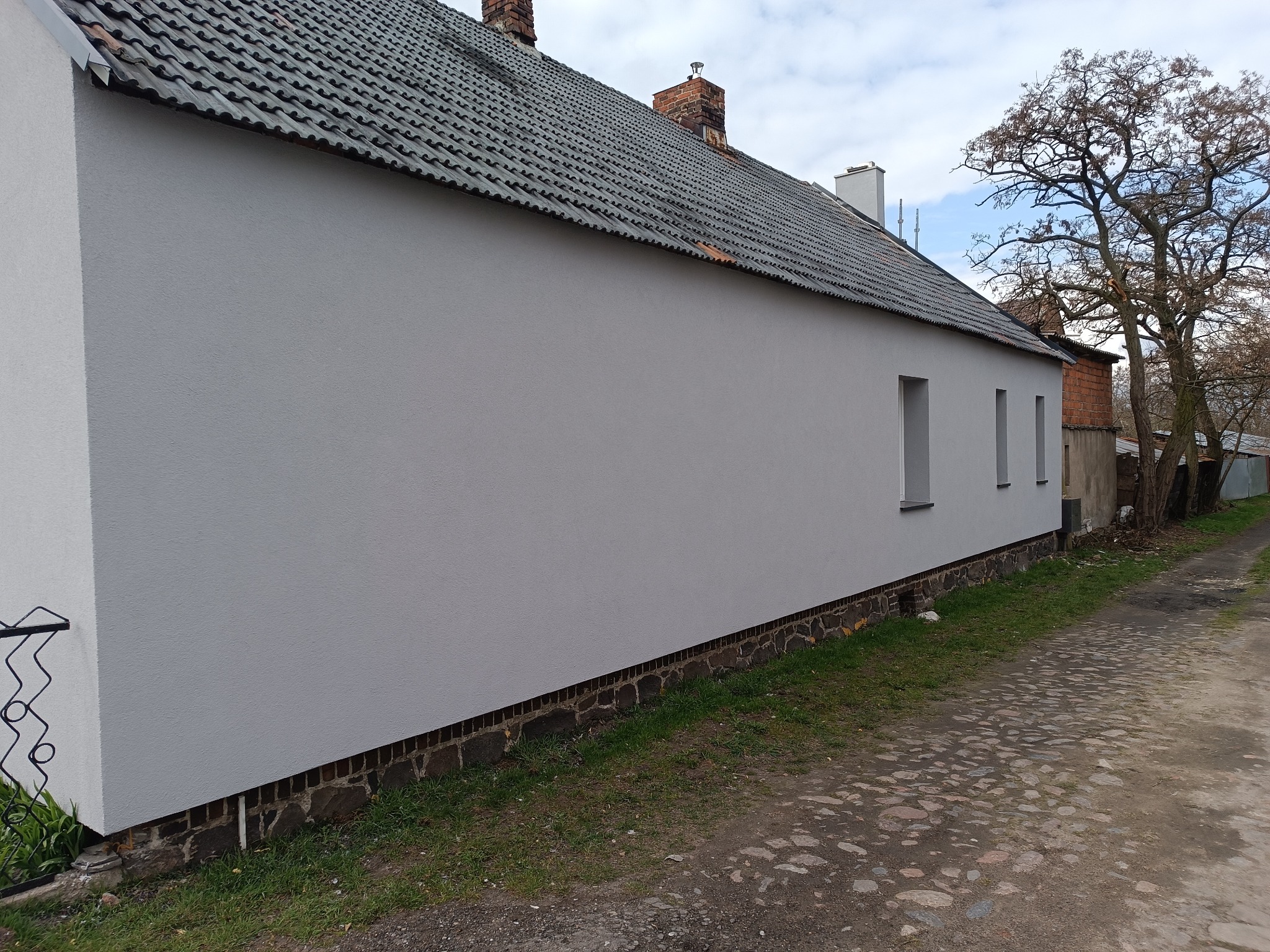 Ściana budynku po ociepleniu i pomalowaniu na szaro, widoczne trzy małe okna, dach z ciemnej dachówki i kamienista droga. Budynek z zewnątrz.