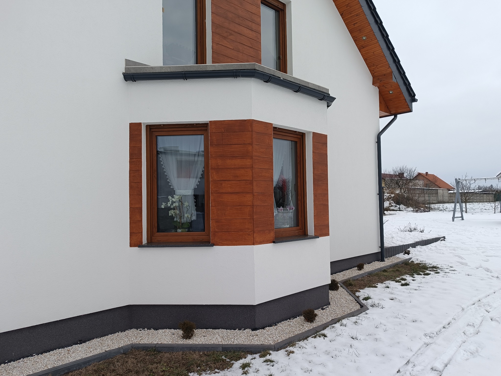 Biała elewacja domu z drewnianymi akcentami okiennymi i wykończeniem cokołu. Śnieżny krajobraz w tle, widoczne rynny i elementy dachu.
