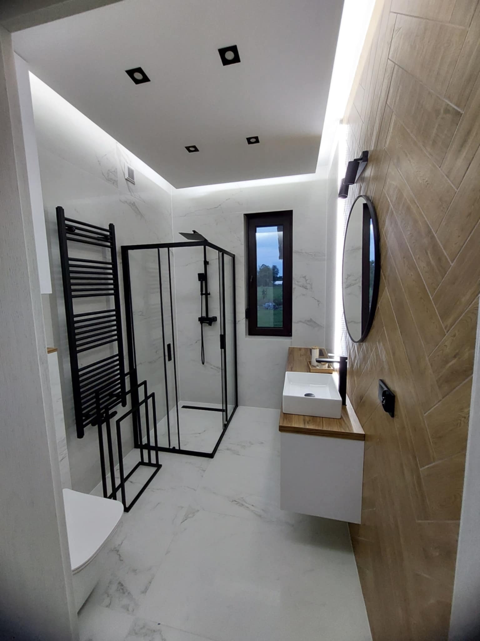 Nowoczesna łazienka z kabiną prysznicową typu walk-in, grzejnikiem drabinkowym i drewnopodobną ścianą ułożoną w jodełkę. Białe marmurowe płytki i podświetlany sufit.