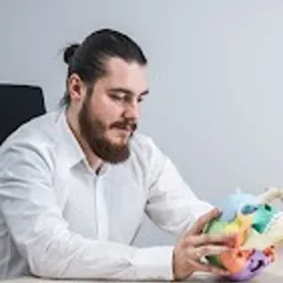 Mężczyzna z brodą i włosami spiętymi w kok, w białej koszuli, siedzi przy biurku i trzyma w dłoniach model kręgosłupa z kolorowymi elementami.