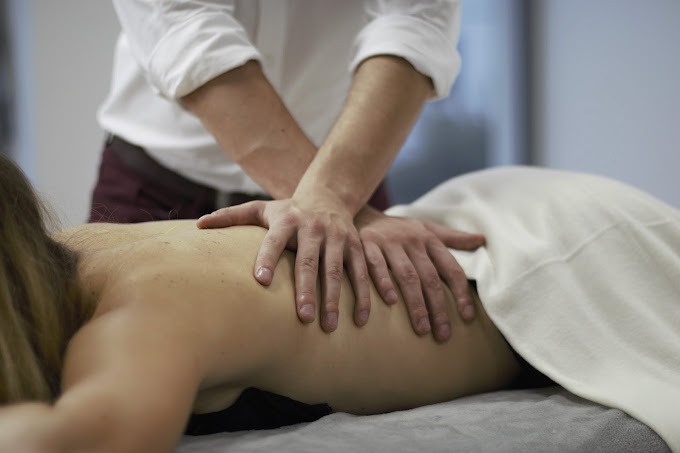 Terapeuta uciska dłońmi plecy pacjentki leżącej na stole pokrytym białym prześcieradłem, widoczne dłonie terapeuty i fragment jego białej koszuli oraz bordowe spodnie.