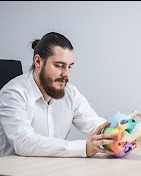 Mężczyzna z brodą i włosami spiętymi w kok, w białej koszuli, siedzi przy biurku i trzyma w dłoniach model kręgosłupa z kolorowymi elementami.