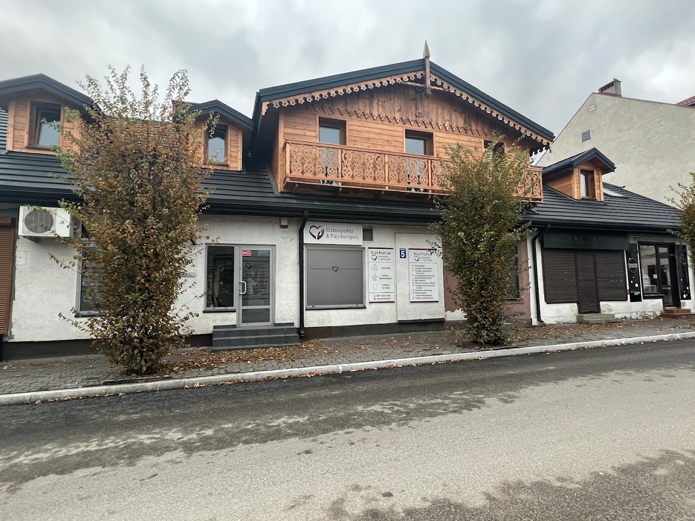 Fasada budynku z szyldem gabinetu z usługami medycznymi, widoczny numer 5, drzwi wejściowe i okna z naklejkami informacyjnymi, stylizowana górna część budynku z drewnianym balkonem.
