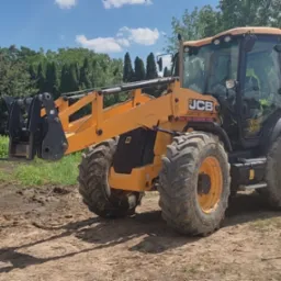 Żółta koparko-ładowarka JCB na placu budowy z widocznymi śladami opon na ziemi i kałużami, w tle zieleń i dom jednorodzinny.