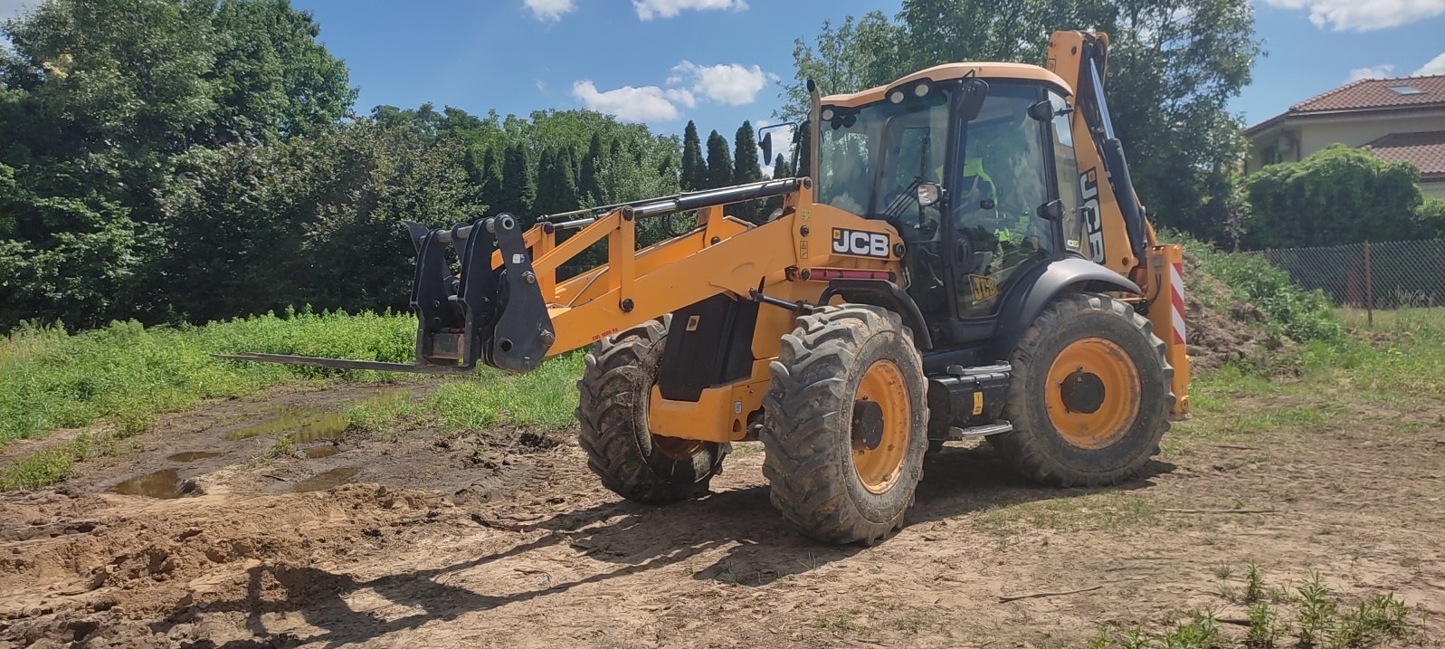 Żółta koparko-ładowarka JCB na placu budowy z widocznymi śladami opon na ziemi i kałużami, w tle zieleń i dom jednorodzinny.