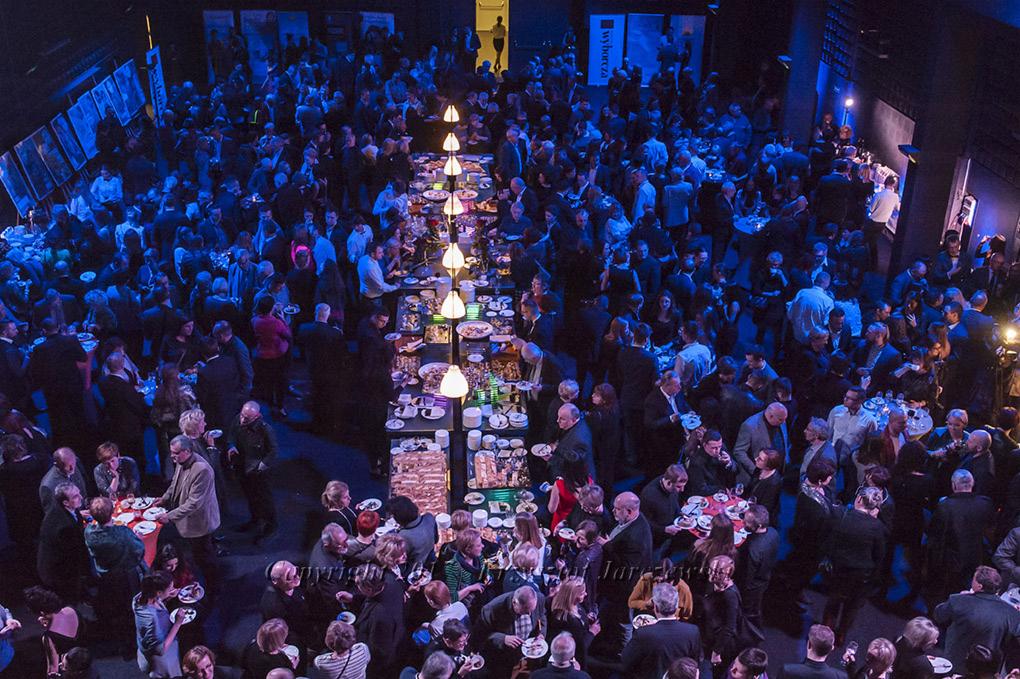 Widok z góry na dużą salę bankietową wypełnioną gośćmi, z długim stołem zastawionym jedzeniem i oświetlonym lampami, w tle ciemne ściany i oświetlenie w odcieniach niebieskiego.