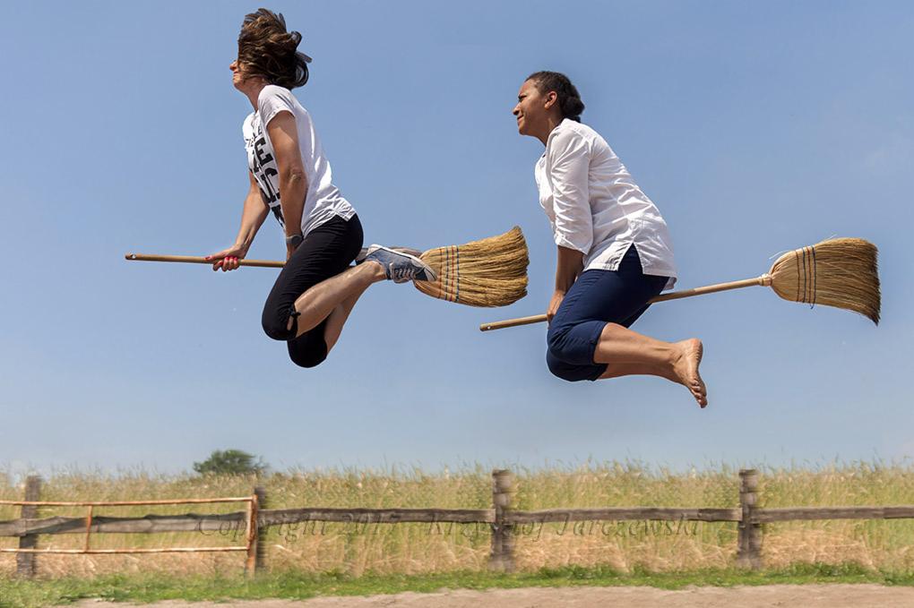 Dwie kobiety w powietrzu, udające lot na miotłach, w plenerze z drewnianym płotem i polem w tle, na tle błękitnego nieba.