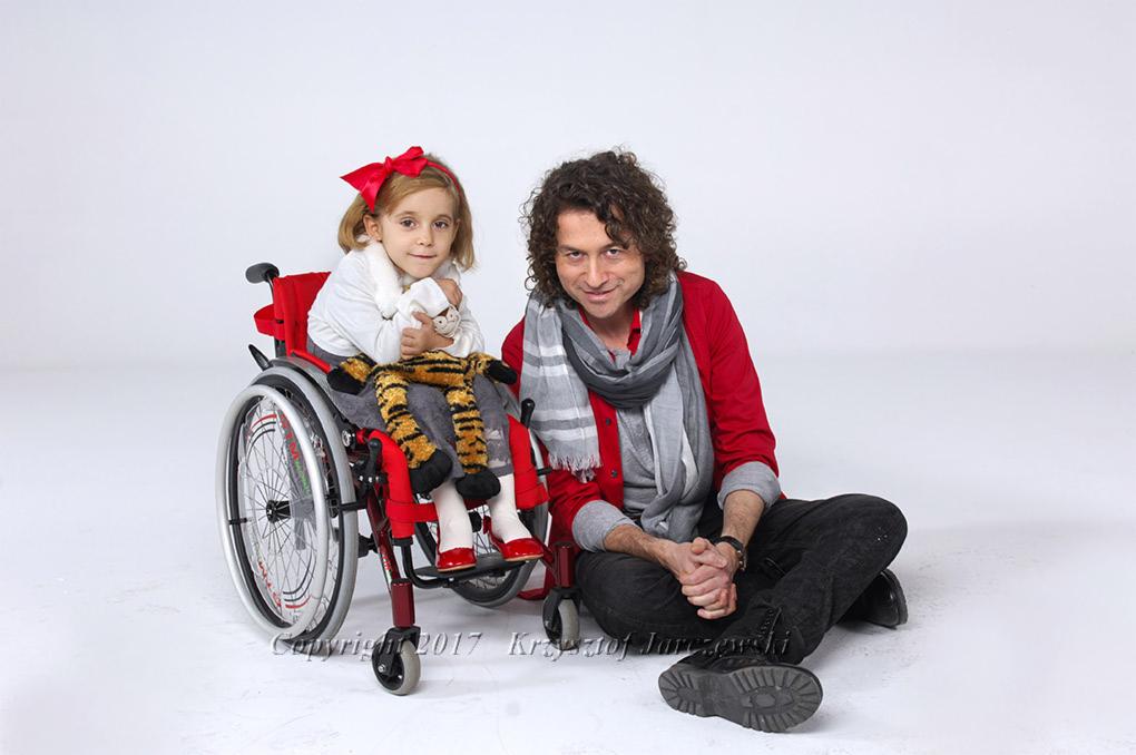 Portret dziewczynki siedzącej na wózku inwalidzkim, trzymającej pluszowego tygrysa, obok siedzi mężczyzna z szalikiem na białym tle.