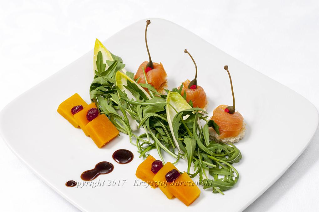 Elegancko podane danie na białym talerzu: plastry pieczonej dyni z żurawiną, roladki z łososia z kaparami na grzance, rukola i kropla balsamicznego sosu.