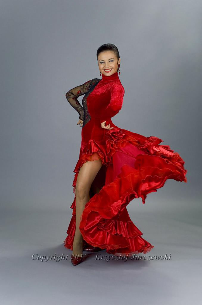 Uśmiechnięta kobieta w czerwonej sukni do tańca z falbanami i czarnymi koronkowymi rękawami pozuje na szarym tle.