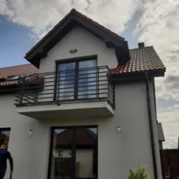 Dom z balkonem z czarną, metalową balustradą o minimalistycznym designie. Widoczne odbicie nieba w szybach okien. Na pierwszym planie roślina w doniczce.