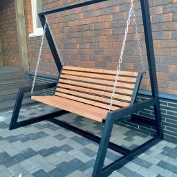 NEXUS Wykopy Kacper Bronk - Metalowa huśtawka ogrodowa z drewnianym siedziskiem, zawieszona na łańcuchach. Nowoczesny design, ciemna rama, jasne drewno, tło to ceglana ściana i kostka brukowa.