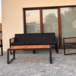 Komplet mebli ogrodowych: sofa trzyosobowa, sofa dwuosobowa i stolik kawowy. Konstrukcja z czarnego metalu, drewniane wstawki, czarne poduszki. Ustawione na szarym bruku.