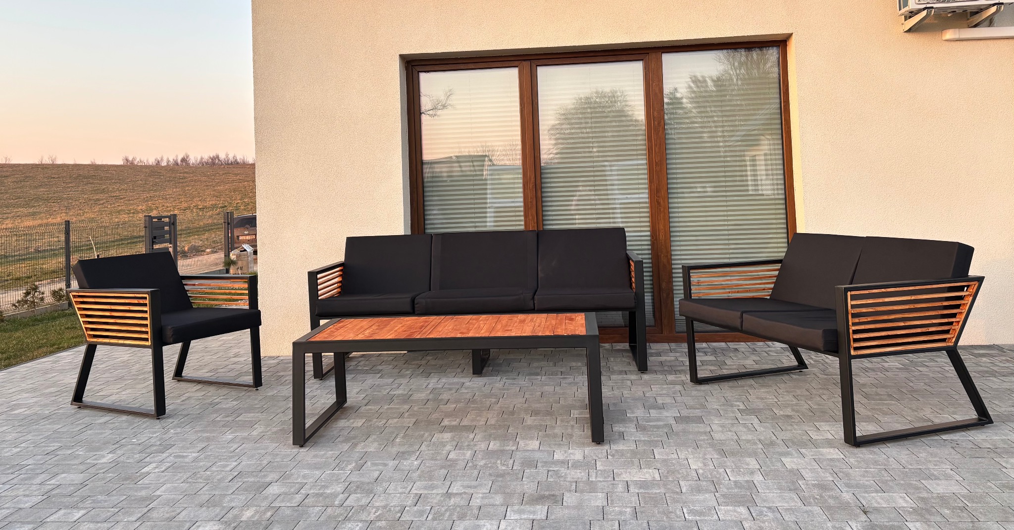 Komplet mebli ogrodowych: sofa trzyosobowa, sofa dwuosobowa i stolik kawowy. Konstrukcja z czarnego metalu, drewniane wstawki, czarne poduszki. Ustawione na szarym bruku.