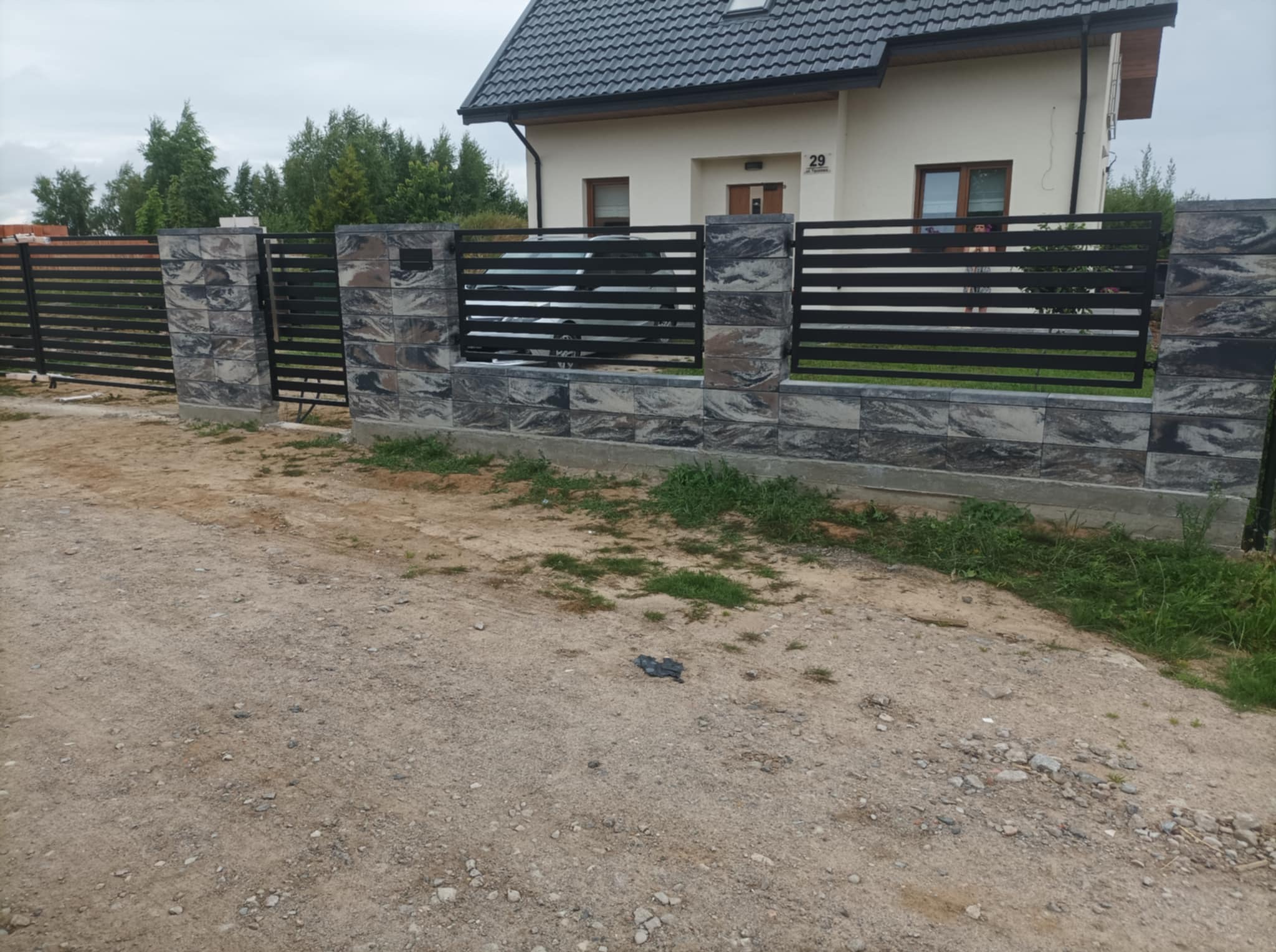 Nowoczesne ogrodzenie posesji z czarnymi, poziomymi przęsłami metalowymi i słupkami z szarej, dekoracyjnej kostki. Dom z kremową elewacją w tle.