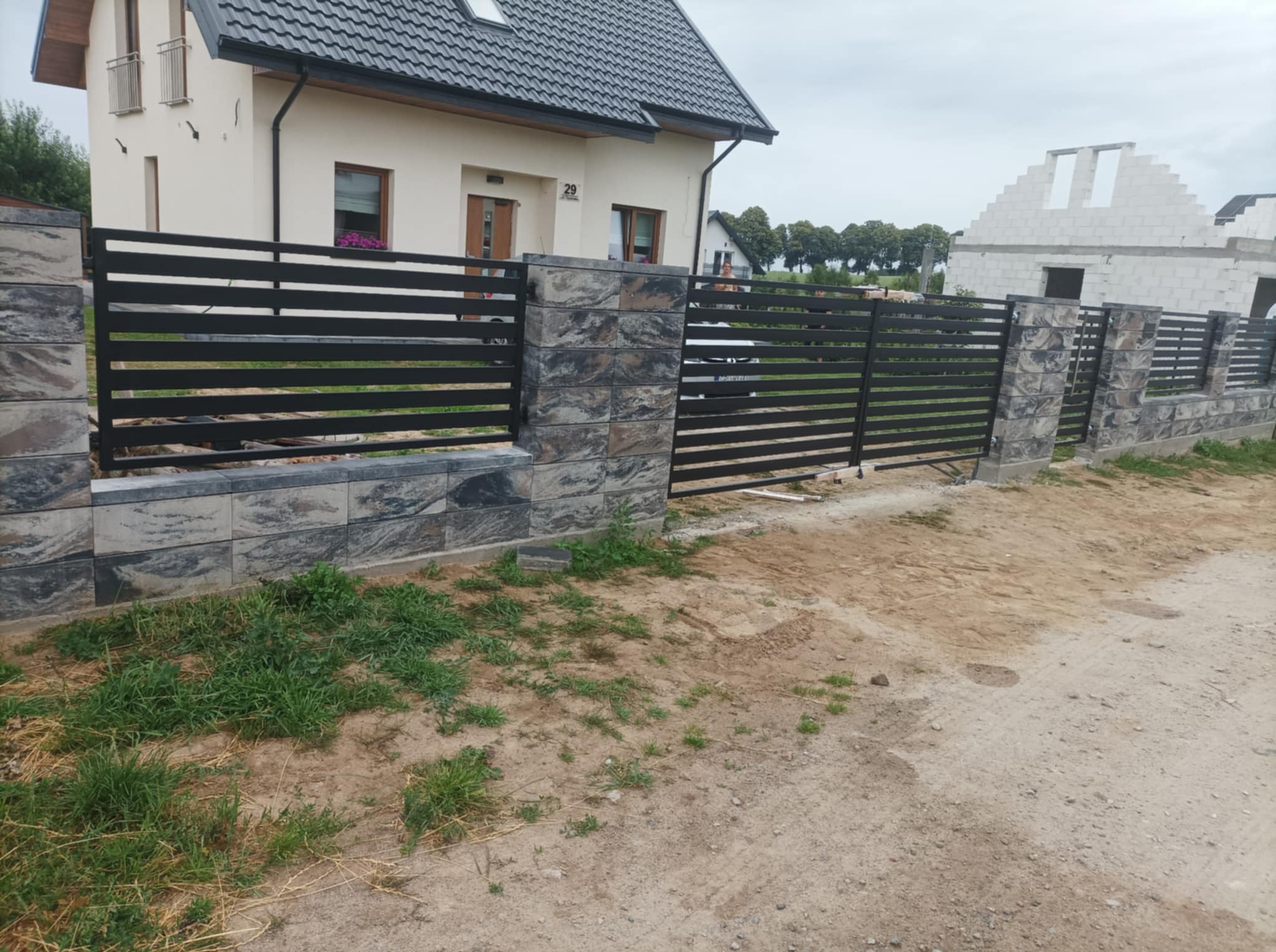 Nowoczesne ogrodzenie posesji z poziomymi, czarnymi przęsłami i kamiennymi słupkami. W tle dom z ciemną dachówką i budowa w stanie surowym otwartym.