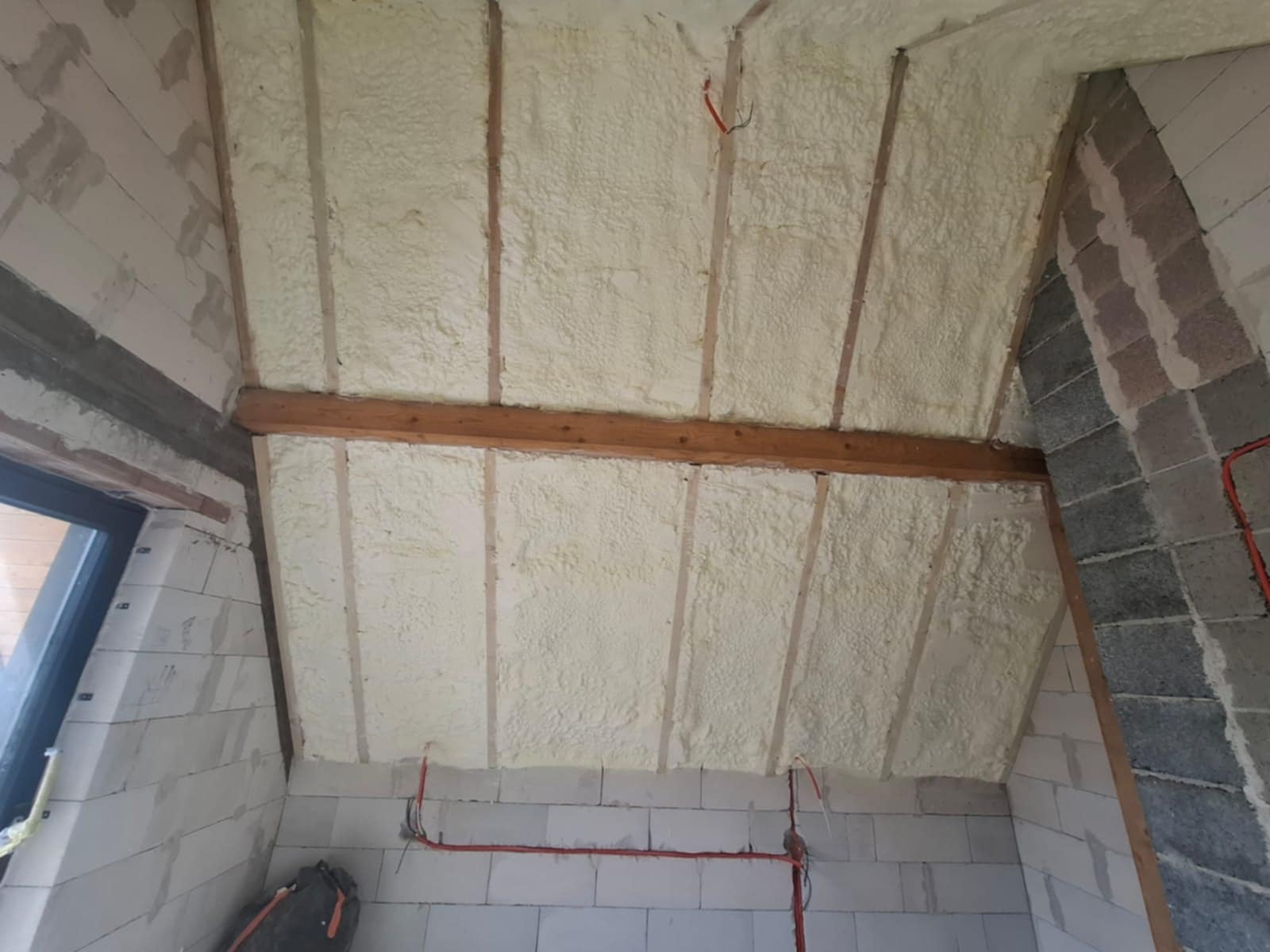 Poddasze z drewnianą konstrukcją stropu pokryte jasną pianką izolacyjną, widoczne przewody elektryczne i fragment ściany z bloczków.