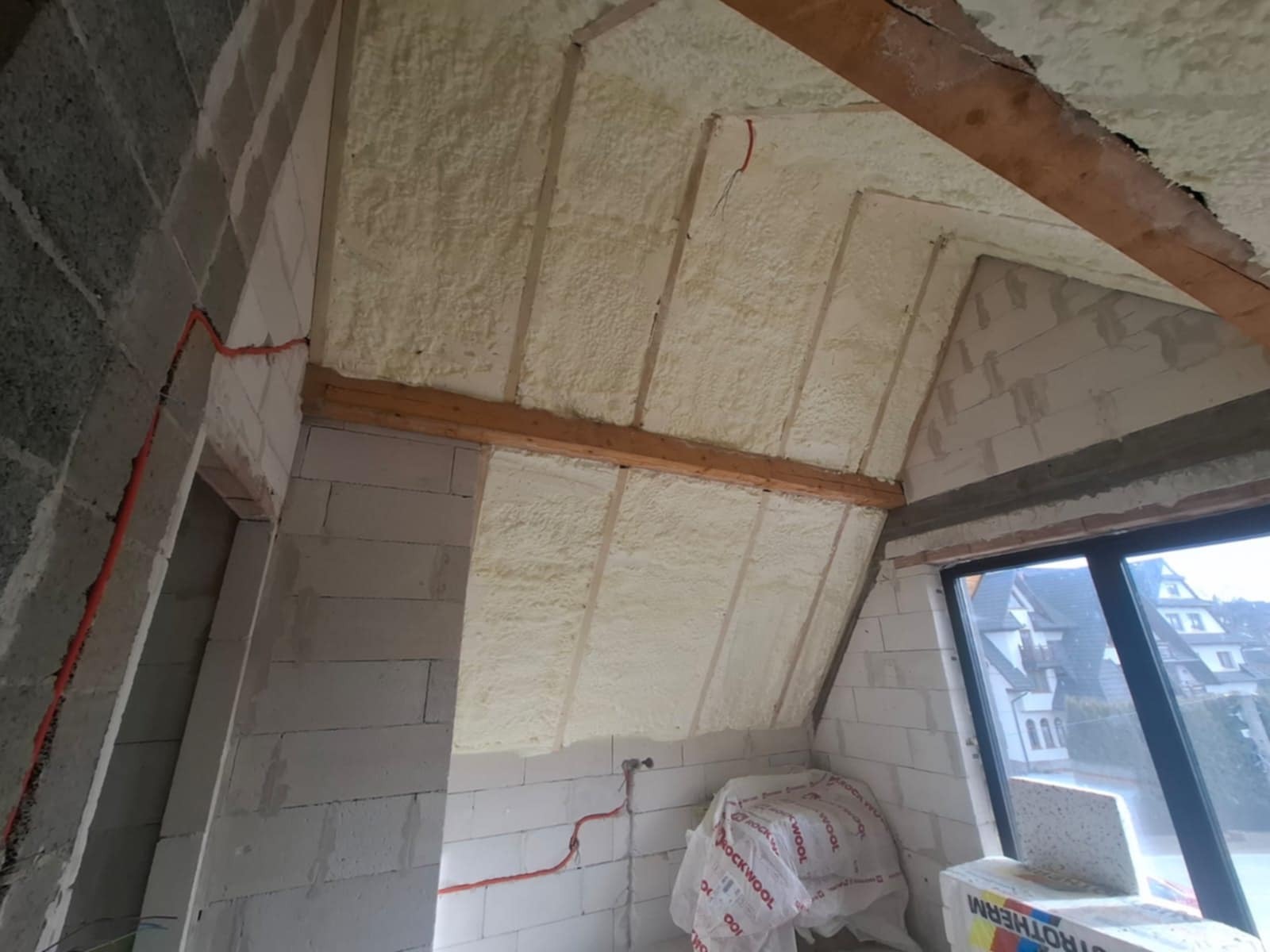 Poddasze w trakcie izolacji pianką poliuretanową, widoczne drewniane belki konstrukcyjne i ściany z bloczków betonowych, częściowo ocieplone, z widocznymi przewodami elektrycznymi.