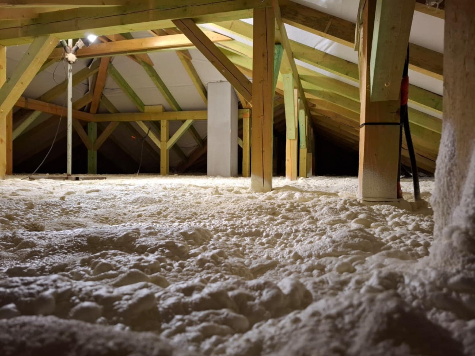 Poddasze z drewnianą konstrukcją pokryte grubą warstwą jasnej, spienionej izolacji, oświetlone lampą roboczą, widoczne słupy i elementy konstrukcyjne.
