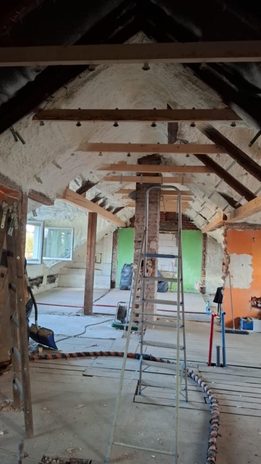 Poddasze w trakcie ocieplania pianką PUR, widoczne drewniane belki i fragmenty muru, drabina, przewody oraz ślady prac budowlanych.