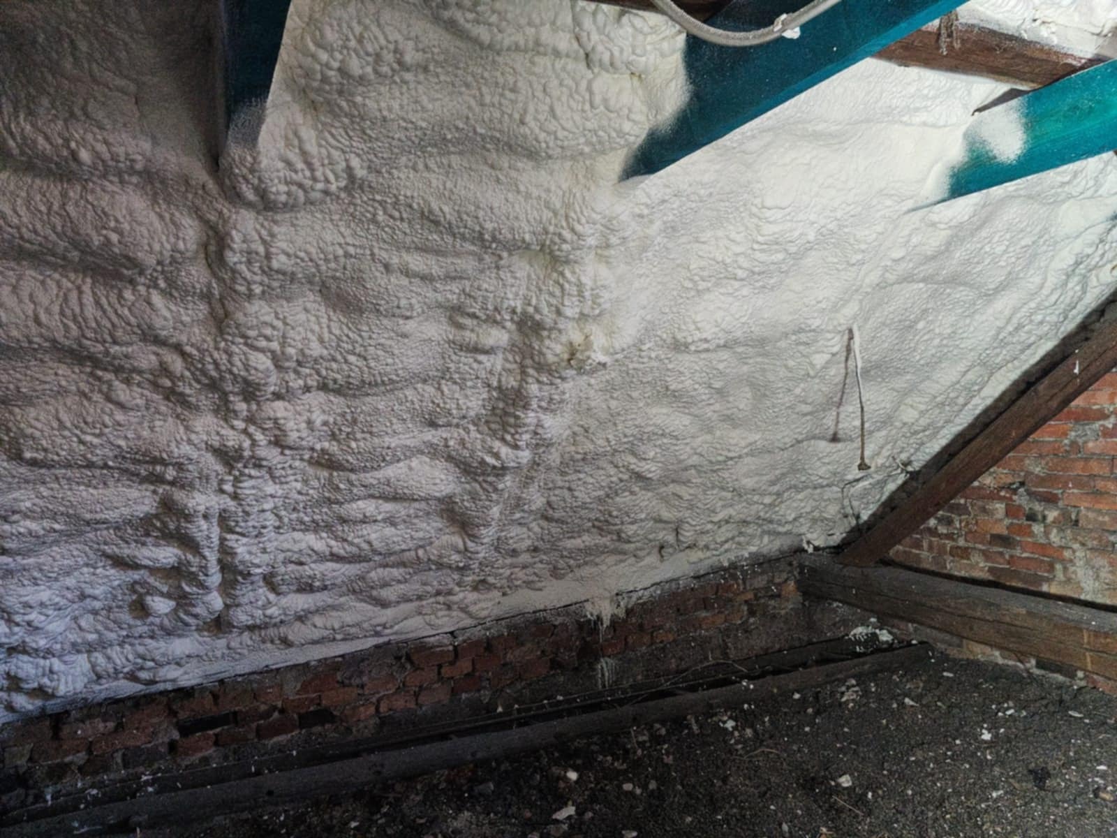 Poddasze z widoczną warstwą izolacyjną z pianki, na tle ceglanej ściany i drewnianych belek konstrukcyjnych, z fragmentem nieocieplonej powierzchni.