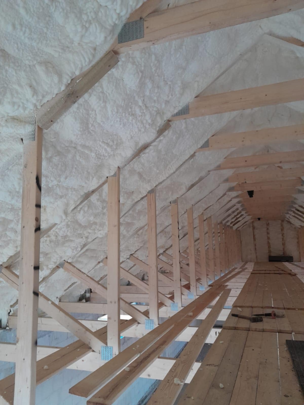 Poddasze w trakcie ocieplania: widoczne drewniane krokwie, metalowe łączniki i warstwa białej pianki izolacyjnej pokrywająca dach, częściowo ułożona podłoga z desek.