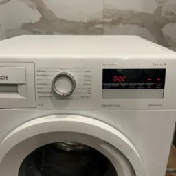 Frontalny widok na białą pralkę Bosch Serie 4 z włączonym panelem sterowania, wskazującym czas prania. Pralka stoi w łazience w Skórczu, na ścianie widoczne płytki imitujące marmur.