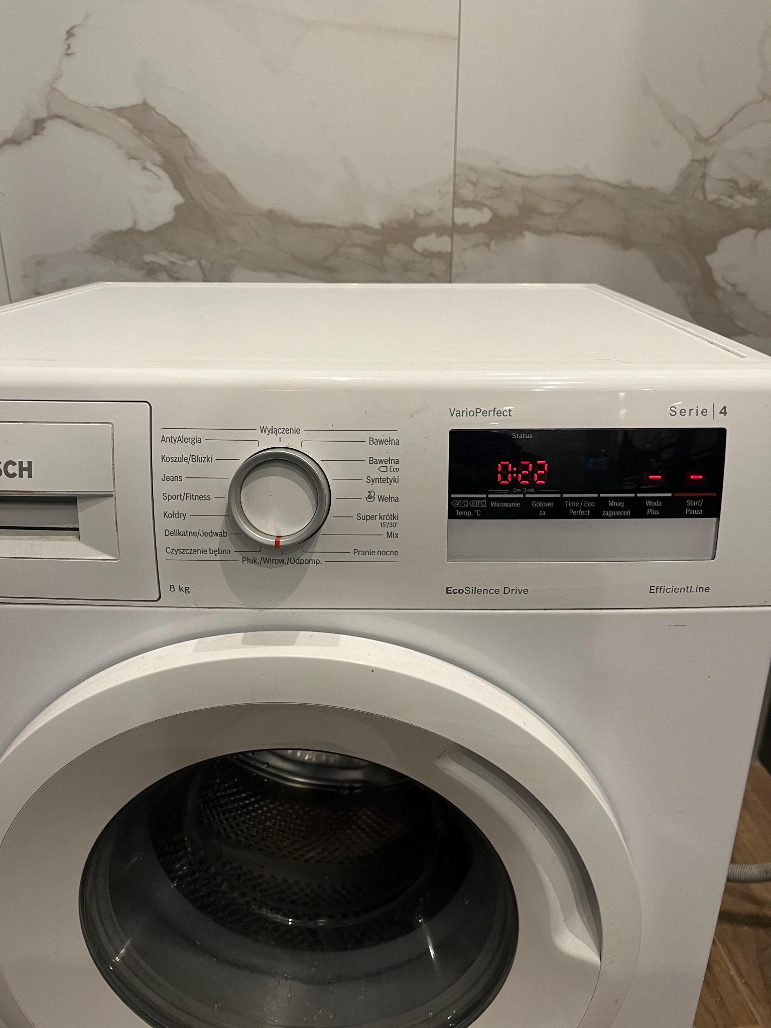 Frontalny widok na białą pralkę Bosch Serie 4 z włączonym panelem sterowania, wskazującym czas prania. Pralka stoi w łazience w Skórczu, na ścianie widoczne płytki imitujące marmur.